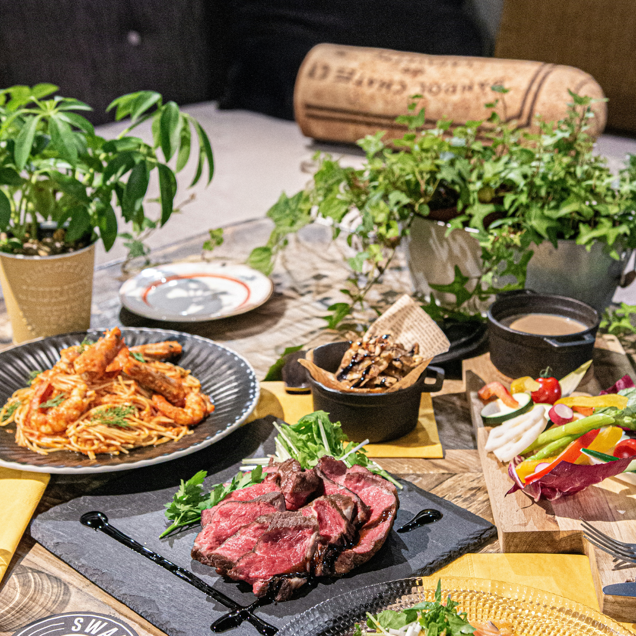 Diverse food platter with steak, pasta, and salad on a table with plants and decor.