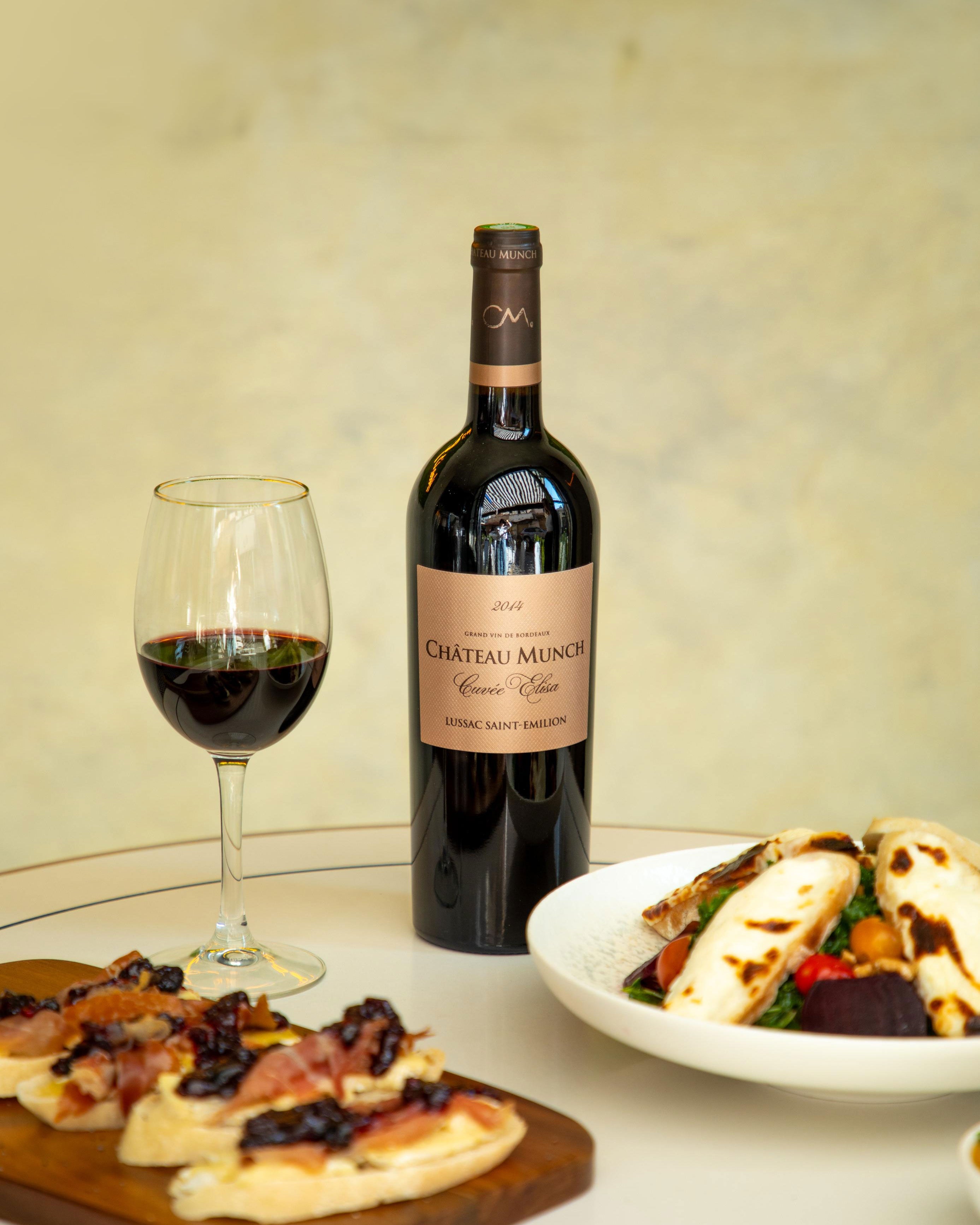 Bottle of Chateau Munch Bordeaux wine with a glass and a plate of food on a table.